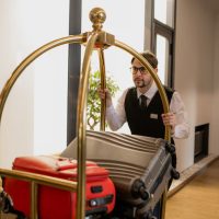 elegant-porter-eyeglasses-pushing-cart-with-pile-suitcases-while-moving-along-corridor-inside-hotel_274679-7092